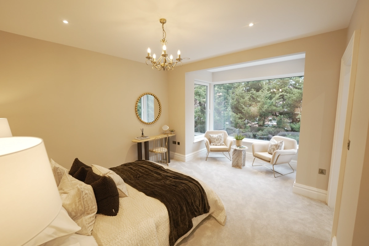 Elegant bedroom with beige walls, featuring a bed with plush pillows and a dark throw. A chandelier hangs from the ceiling. Large bay windows reveal a lush, green outdoor view. Two armchairs and a small table with a plant sit by the window, creating a cozy sitting area.