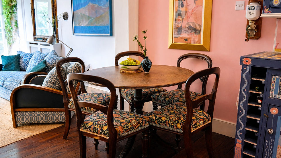 Colorful dining nook with round wooden table, patterned chairs, blue sofa, pink wall, and decorative cabinet; cozy, artistic mood.