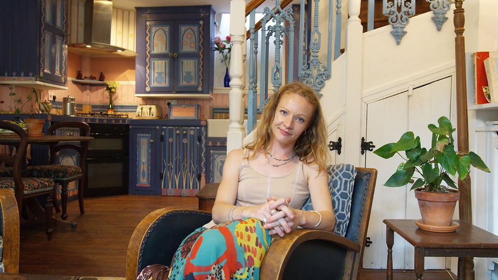 Smiling woman sits in a chair in a colorful vintage kitchen, with blue cabinets, a staircase, and a potted plant nearby.
