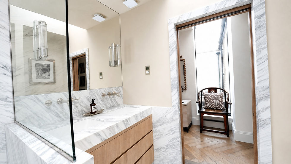 Marble bathroom vanity with mirror and soap dispenser, opening to a bright nook with a cushioned wooden chair and herringbone floor.