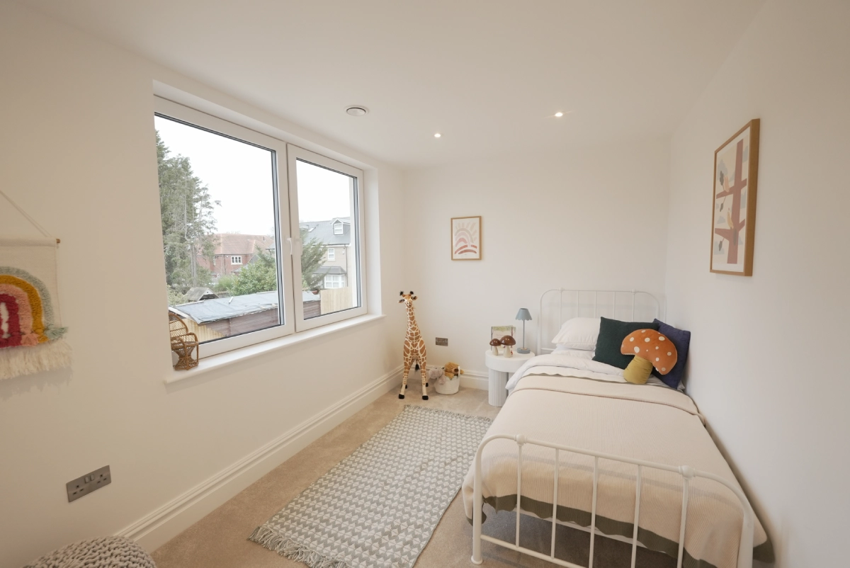 Bright children's bedroom with a single bed, giraffe toy, and colorful decor. Large window offers natural light. Walls are white; room feels cozy.