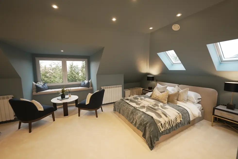 Cozy bedroom with a double bed, pillows, and throw blanket. Skylights and window view. Two chairs, table, and plants create a serene mood.