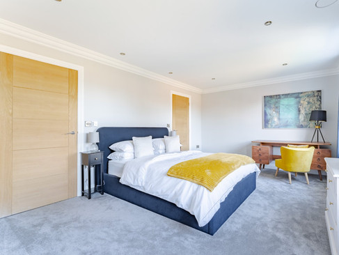 Contemporary bedroom with navy upholstered bed, yellow throw, grey carpet and a wooden desk beneath abstract wall art.
