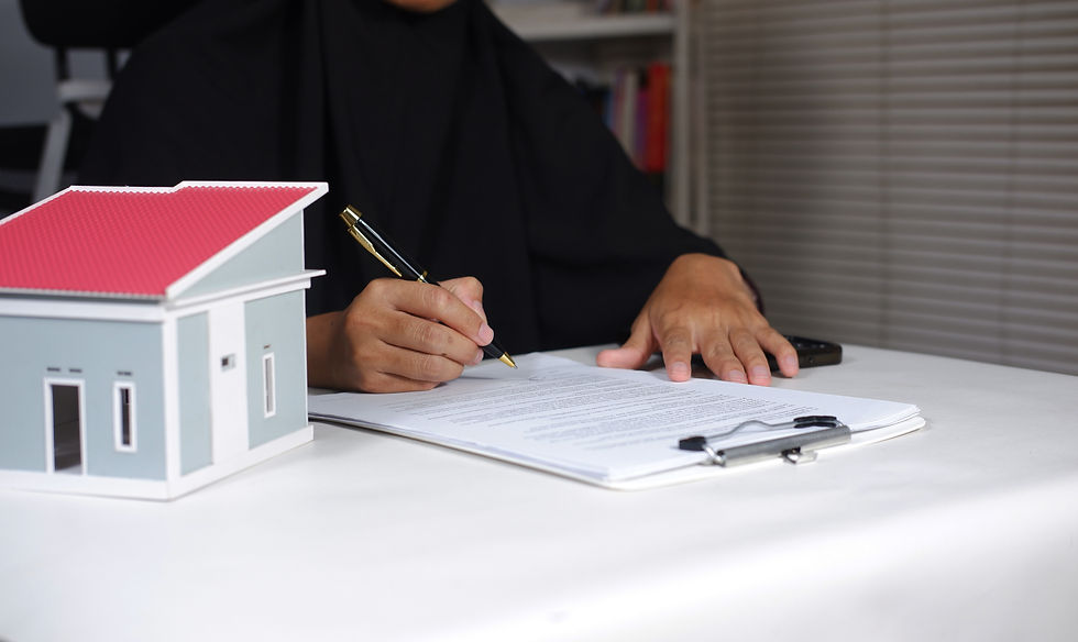 A person signing documents at a table with a small model house nearby. The background includes a bookcase and blinds. The mood is focused.