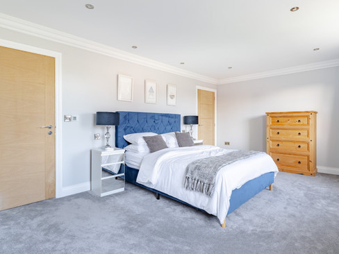 Modern bedroom with deep blue headboard, white bedding, grey carpet and a wooden chest of drawers against pale grey walls.