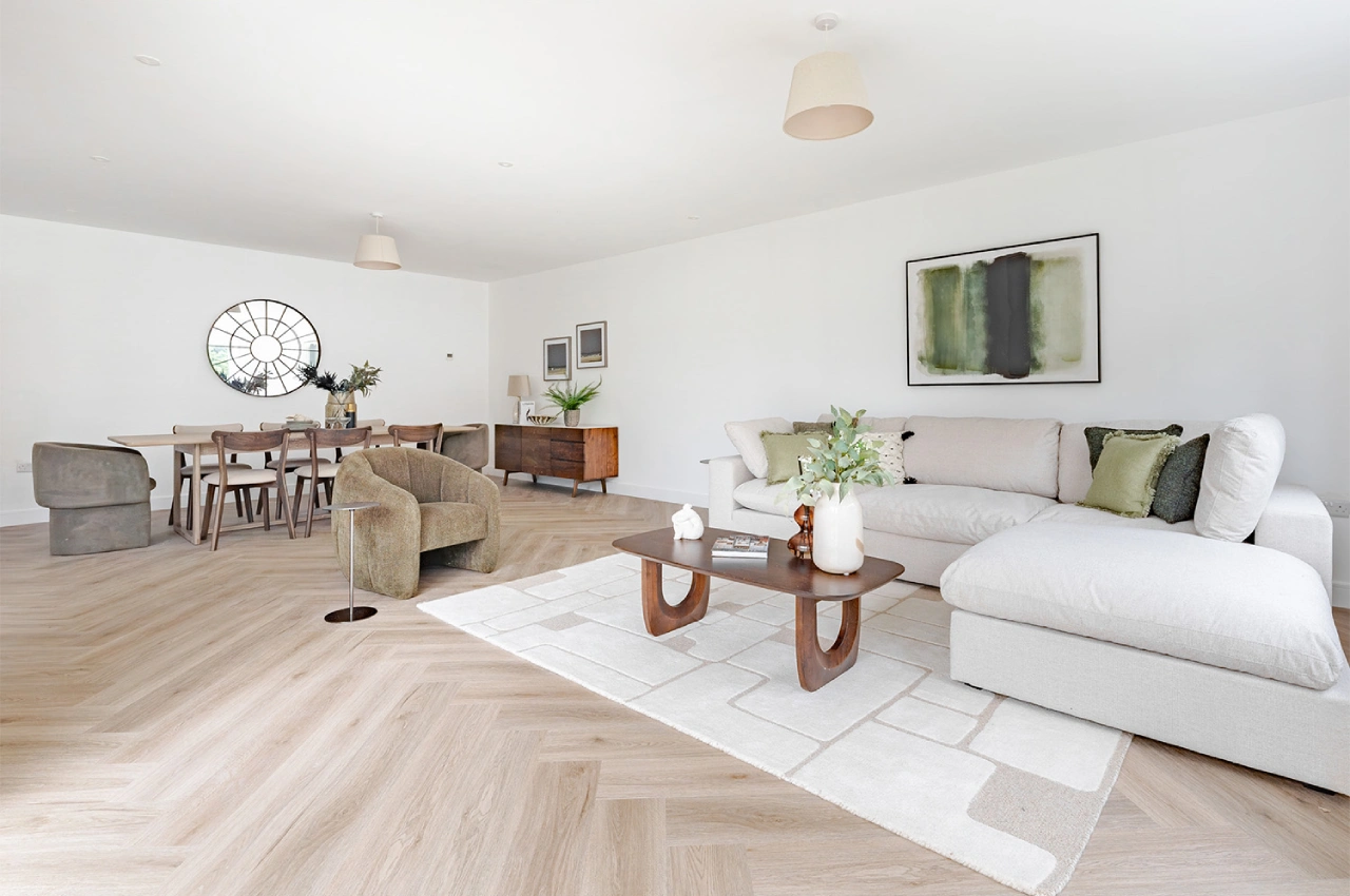 Spacious living room with a light wood herringbone floor, featuring a large beige sectional, green cushions, modern art, and a wood dining set under a round window.
