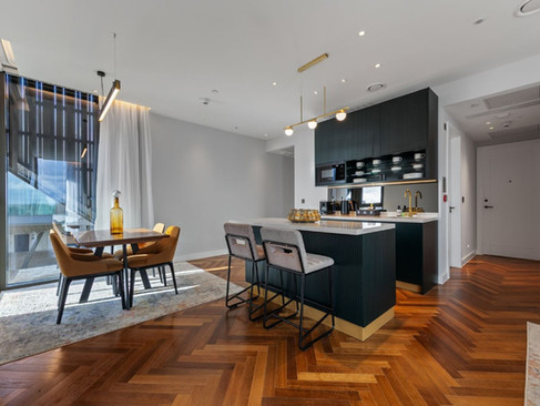 Contemporary kitchen with fluted teal cabinetry, white worktops, brass lighting, dining table with tan chairs and warm parquet flooring.