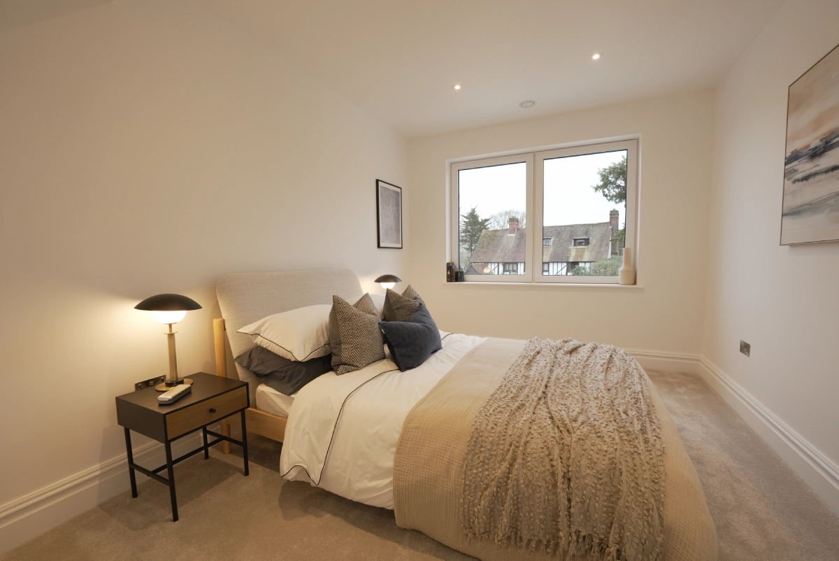 Cozy bedroom with a neatly made bed, soft neutral colors, and decorative pillows. A side table lamp casts a warm glow. Window shows a suburban view.