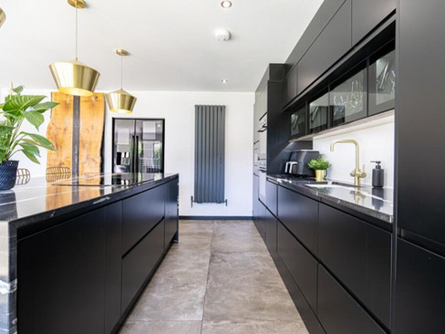 Sleek contemporary kitchen with matte black cabinetry, large island with marble-style worktop, brass pendant lights and floor-to-ceiling glazing at the far end.