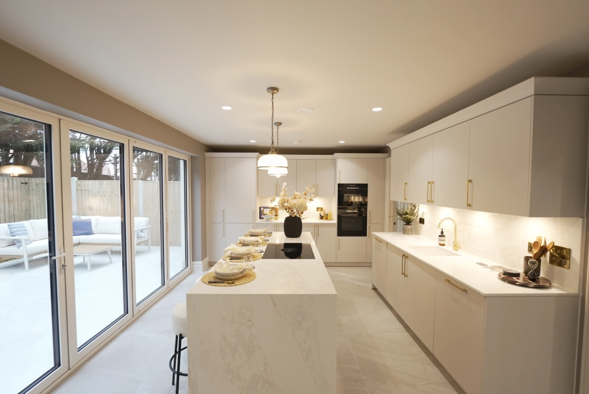 Sleek modern kitchen with white cabinets, large marble island set for dining, under pendant lighting. Glass doors reveal a patio with outdoor seating.