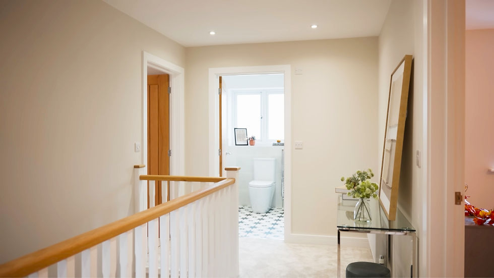 Cream hallway with wooden railing leads to a bathroom. A glass table holds a vase of flowers. Black and white floor tiles visible. Calm atmosphere.