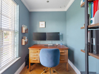 Home office with wooden desk, dual monitors, blue accent walls, window blinds and built-in shelving for storage.