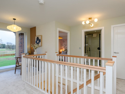 A bright hallway with beige carpet features a wooden handrail, doors to rooms, soft lighting, framed art, and floor-to-ceiling windows overlooking a lawn.