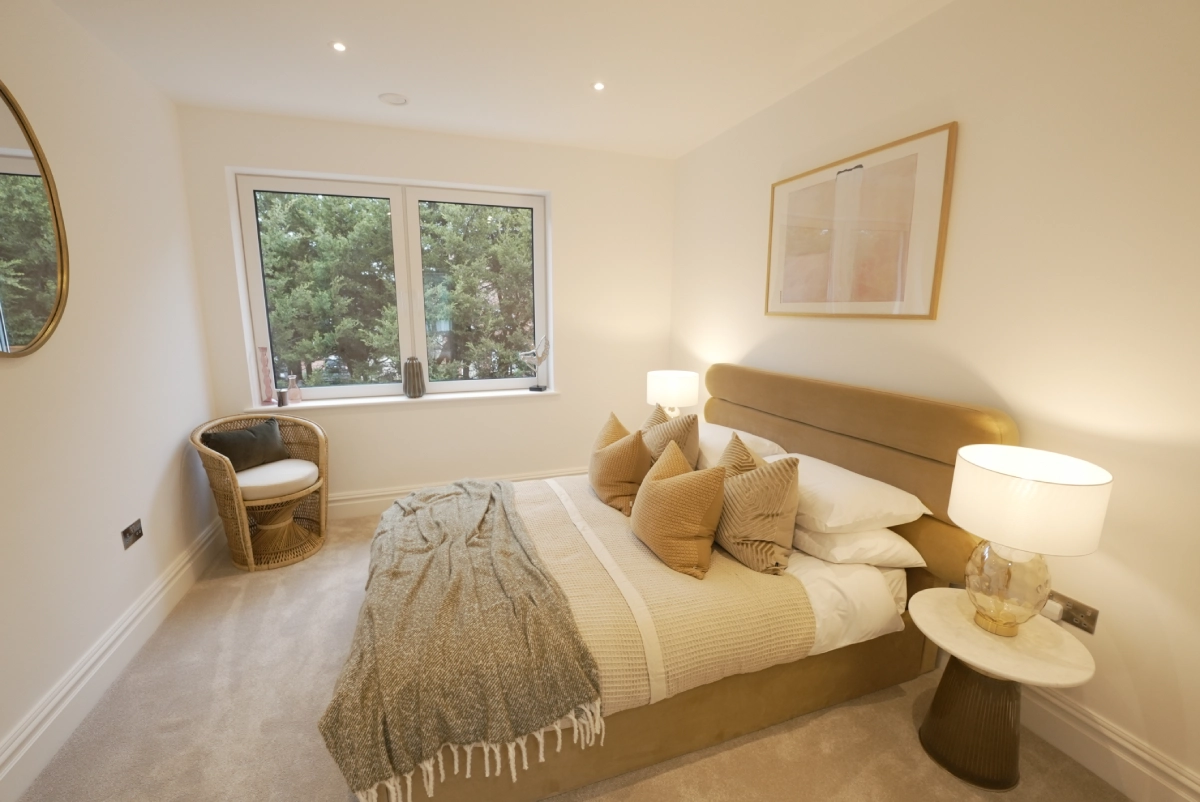 A cozy bedroom with a beige palette features a neatly made bed with a taupe headboard, plush pillows, warm lighting from two table lamps, and a window view of trees.