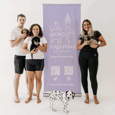 A group of three people poses in front of a Yoga Paws banner, holding puppies while a Dalmatian puppy stands in front.