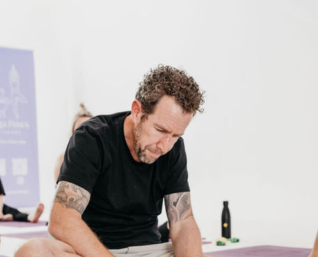 A bearded man in a black shirt stretches on a yoga mat while a small puppy plays beside him.