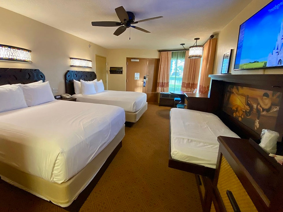 Hotel room with two queen beds, folded wall bed, and a mounted TV showing a castle. Warm lighting, brown patterned carpet, cozy mood.
