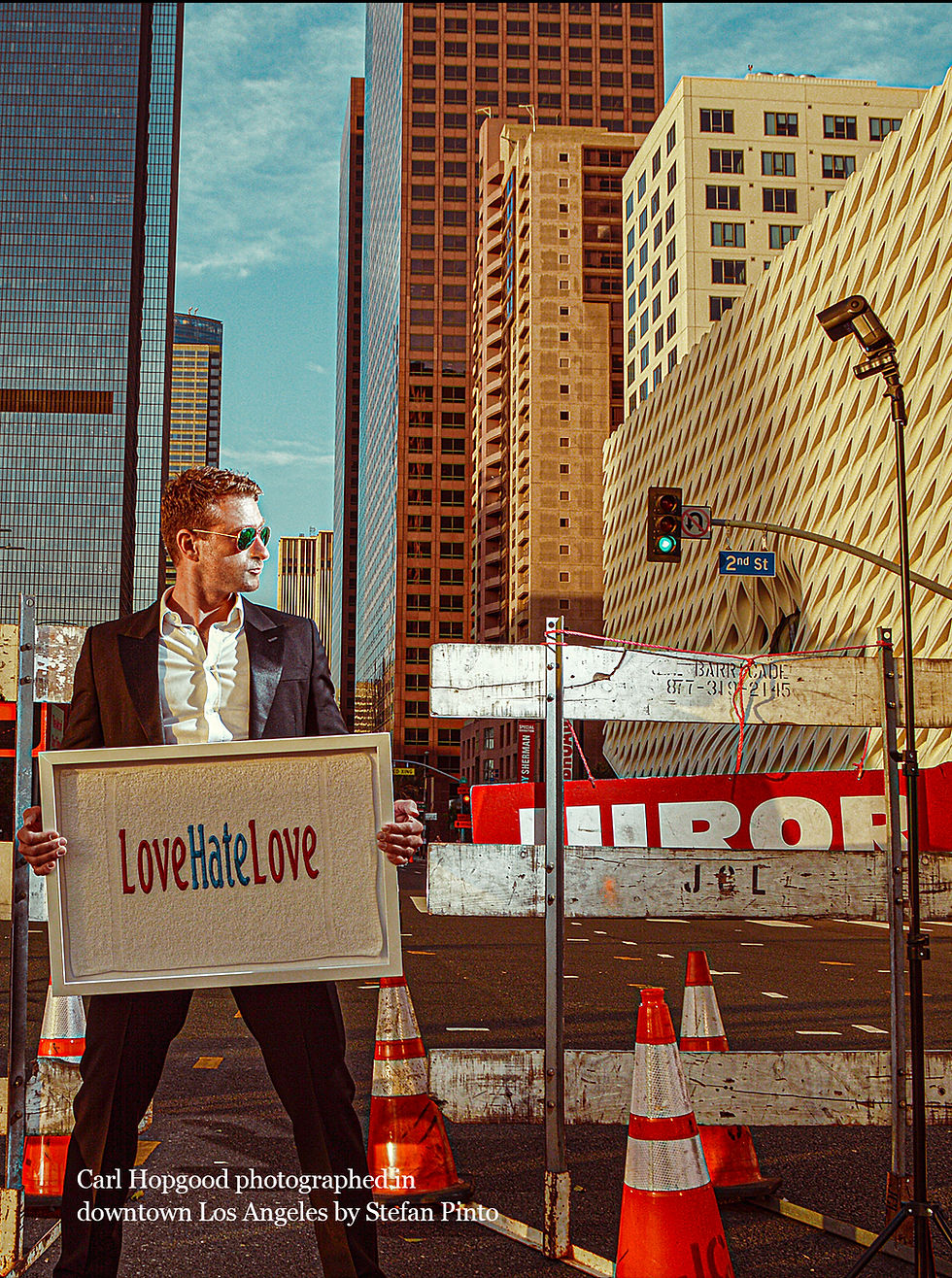 Carl Hopgood, photographed by Stefan Pinto, stands in downtown Los Angeles wearing a black suit, white shirt, and aviator sunglasses. He holds a framed artwork reading “LoveHateLove” in red and blue letters. Behind him, tall skyscrapers, construction barriers, traffic cones, and a 2nd Street sign create an urban backdrop.