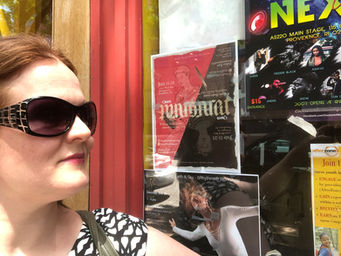 Charlene is wearing sunglasses, looking at a window with a number of signs in it, including one for a performance of the play "Queen Margaret."