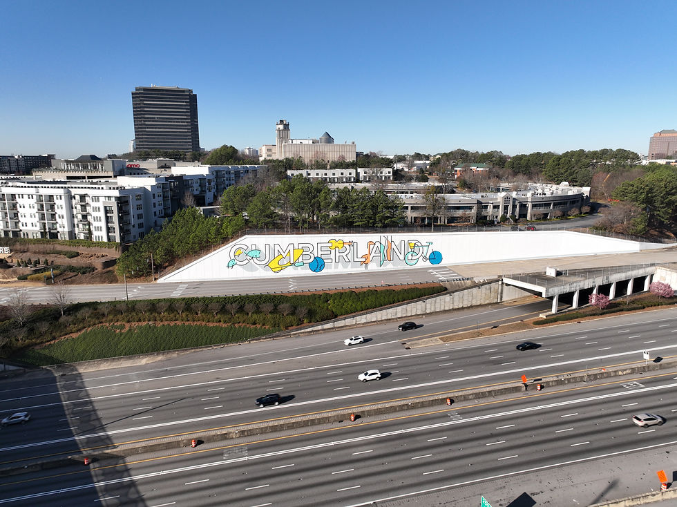 Aerial view of a highway with cars, a mural with "CUMBERLAND" in colorful letters, buildings, and trees under a clear blue sky.