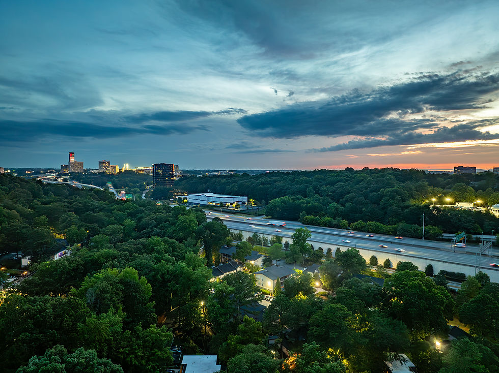 Aerial view of a city at dusk with a highway, buildings, and lush trees. The sky is vibrant with clouds and distant lights glow warmly.