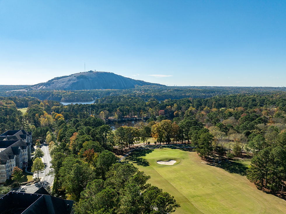 Arial view of a golf course with a sand trap, surrounded by trees in autumn colors. Stone Mountain and blue sky in the background.
