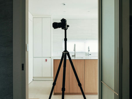 Camera tripod standing in a modern kitchen. White cabinets, wooden accents, and soft lighting create a minimalist and calm atmosphere.