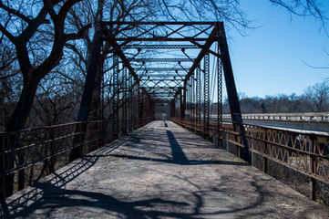 A long, rusty iron bridge at Brazos Point, Texas, stretching toward the horizon with bare trees and a blue sky in the background.
