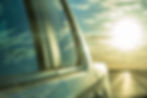 A bright sun low in the sky over a highway, with a green-tinted reflection of blue sky and clouds in a truck's side window.