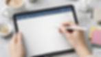 Hands hold a tablet with a digital notepad app, writing with a stylus. A coffee cup, earbuds, and notepad are on a gray table.