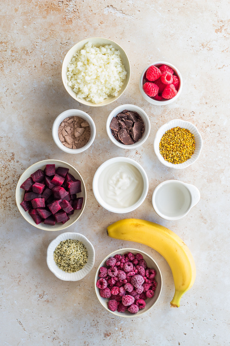 Aerial view of colorful smoothie ingredients: raspberries, chocolate, yogurt, banana, beets, cauliflower, and seeds on a speckled surface.