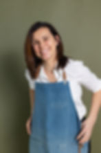 Smiling woman in a blue apron and white shirt stands against a green background. She exudes a cheerful and approachable demeanor.