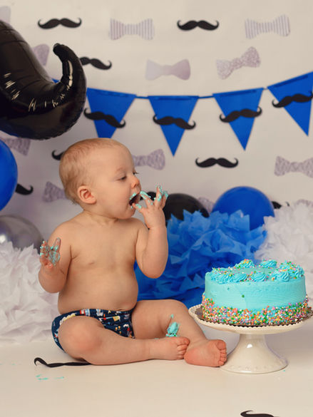 Baby boy with mustache balloons enjoying a blue frosted cake at a mustache-themed cake smash, photographed by Maria Sheehan Photography