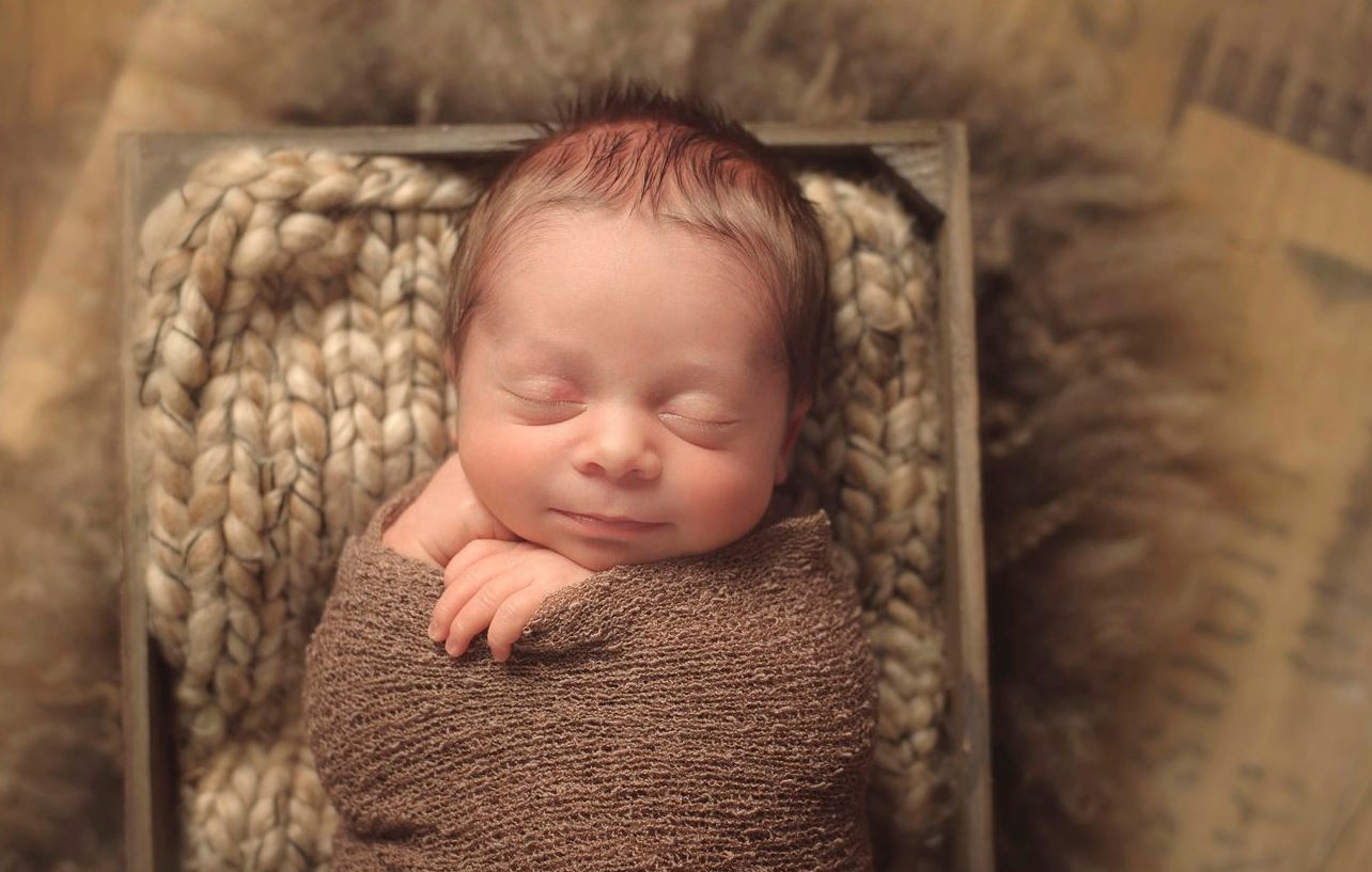 A sleeping newborn wrapped in a brown knit fabric, with a blissful smile, nestled inside a rustic wooden frame against a chunky knit background. The photograph, taken by Maria Sheehan Photography, captures the essence of tranquil newborn slumber and is characteristic of her work in Vancouver, Washington, and Portland, Oregon.