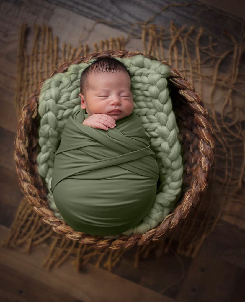 A newborn baby is swaddled in a cozy olive green wrap, nestled within a circular wicker basket lined with a soft, mint green knit blanket, capturing the essence of a natural and serene newborn photography environment.