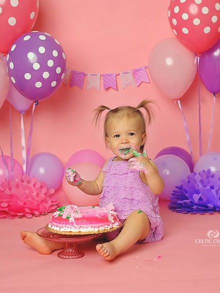 Joyful toddler in a purple dress with a pink polka dot cake smash setup, complete with pastel balloons and paper fans, by Maria Sheehan Photography.