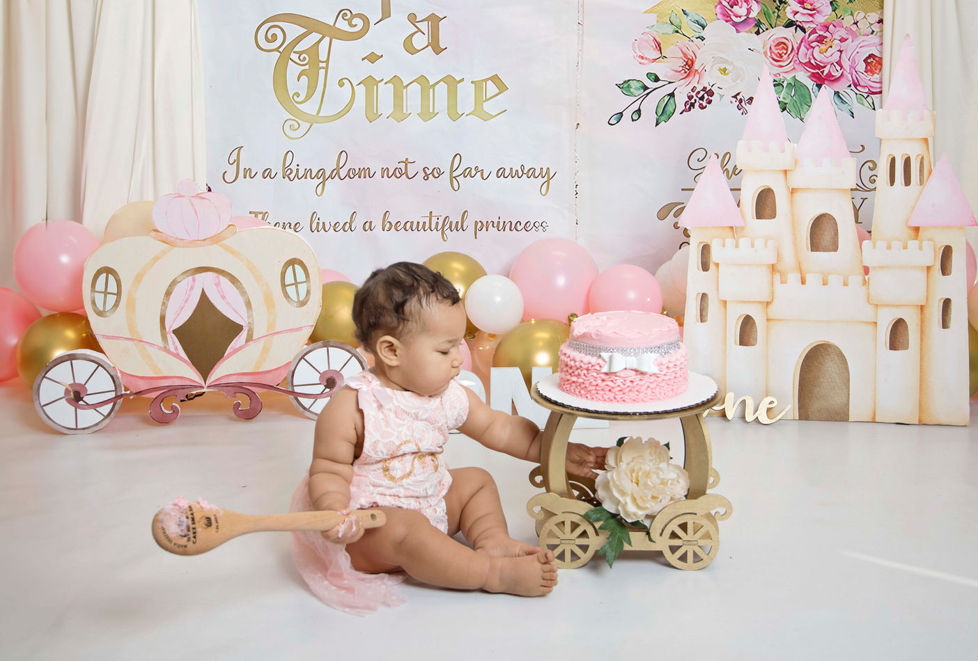 A toddler in a princess-themed cake smash photography session by Maria Sheehan Photography, Vancouver WA, sitting beside a pink frosted cake on a wooden carriage, with a fairytale castle backdrop, surrounded by balloons and a 'Once Upon a Time' banner