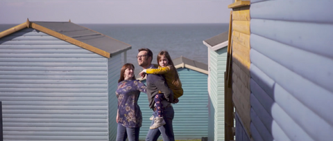 Parents walking through beach huts with their daughter on dad’s shoulders on a sunny day.
