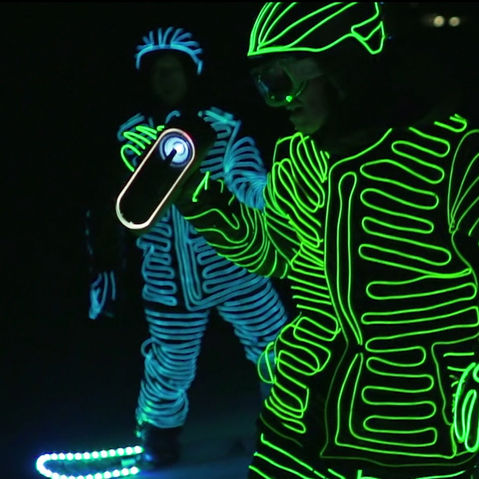Two snowboarders in LED suits holding a Sony speaker as they ride down a snowy slope at night.