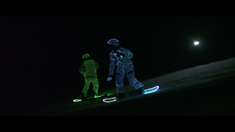 Two snowboarders in LED suits holding a Sony speaker as they ride down a snowy slope at night.