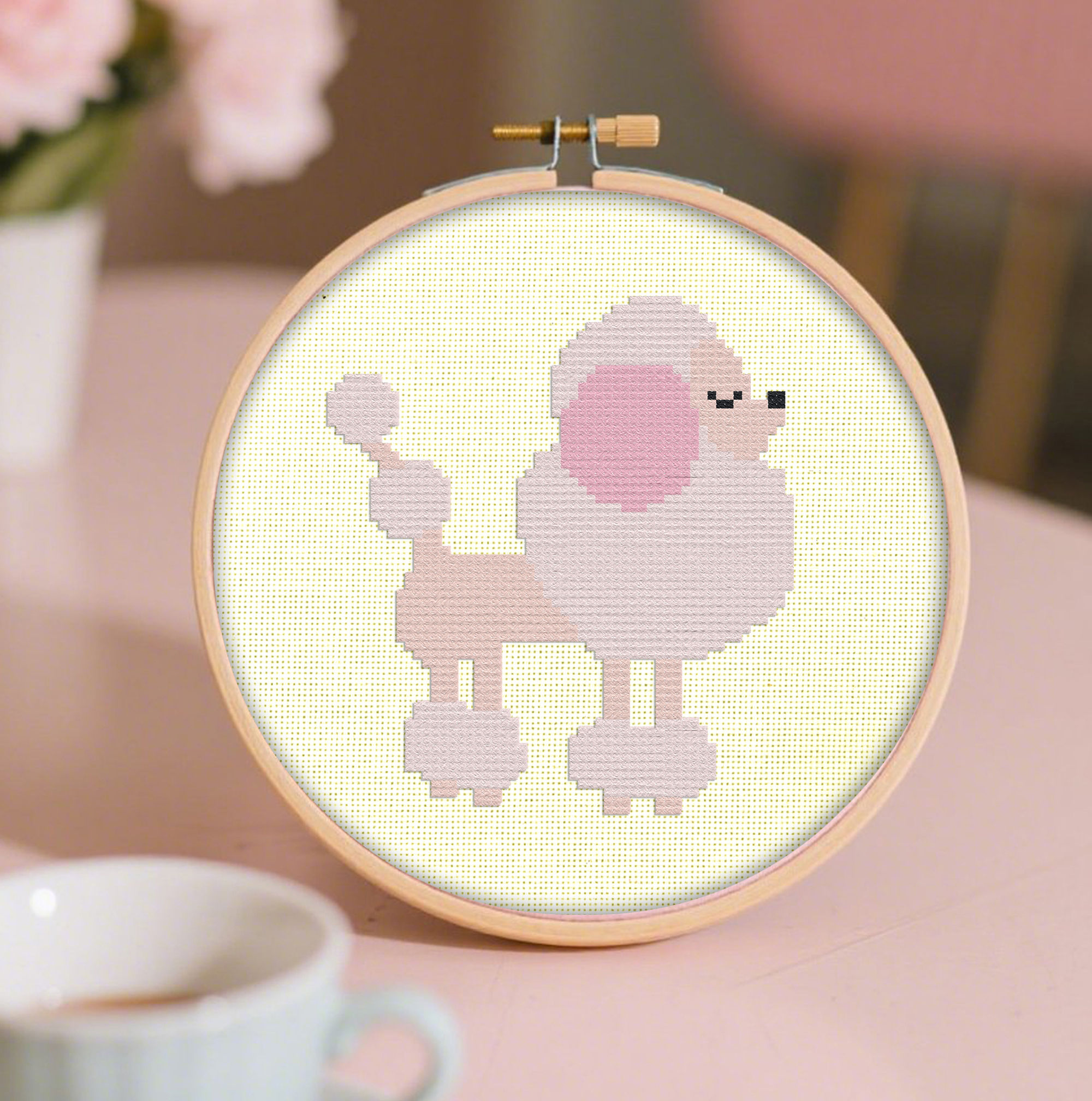 a cross stitch of a retro, pink French poodle displayed in an embroidery hoop next to flowers and a cup of tea