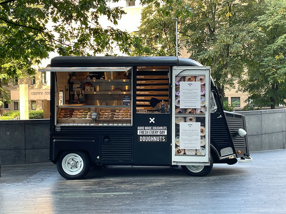 Black doughnut van in a leafy setting, displaying pastries inside. Signs read "Hand Made Doughnuts" and "Card Only." A cozy, inviting vibe.