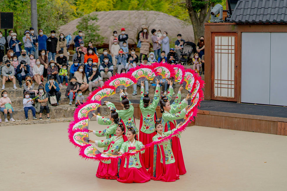 Women in green and red hanbok form a fan circle dance before a masked crowd at an outdoor stage.