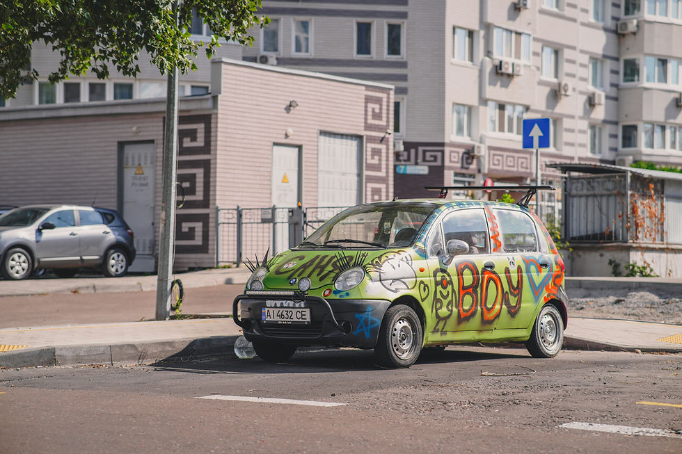 Graffiti-covered green BMW mini car parked on a city street by apartment buildings, with a blue one-way sign.