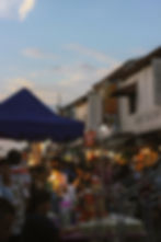 Crowded night market with stalls under blue tent, people browsing items, warm lights creating a cozy atmosphere, shops in background.