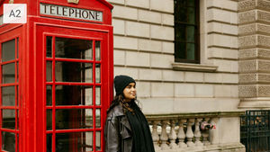 Woman in black beanie and coat stands by a red telephone booth labeled "TELEPHONE." Beige building background, calm expression.