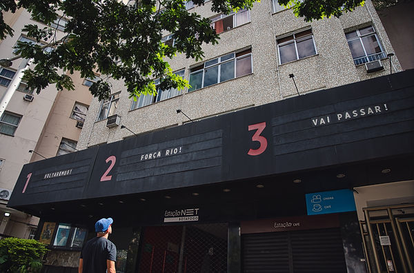 Em frente ao Cinema fechado, um rapaz de boné e blusa azul olha para os letreiros no alto, que dizem: Voltaremos!, Força Rio e Vai passar!