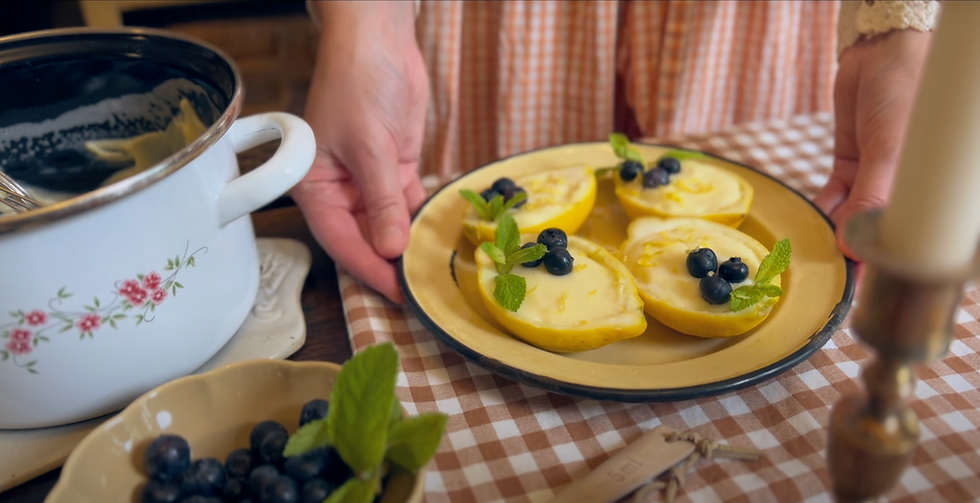 Hands holding a plate of lemon desserts with blueberries and mint, on a brown checkered tablecloth beside a floral pot and candle.