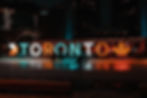 Skaters glide on an ice rink at night in front of lit "Toronto" sign with festive lights. Dark city skyline in the background.