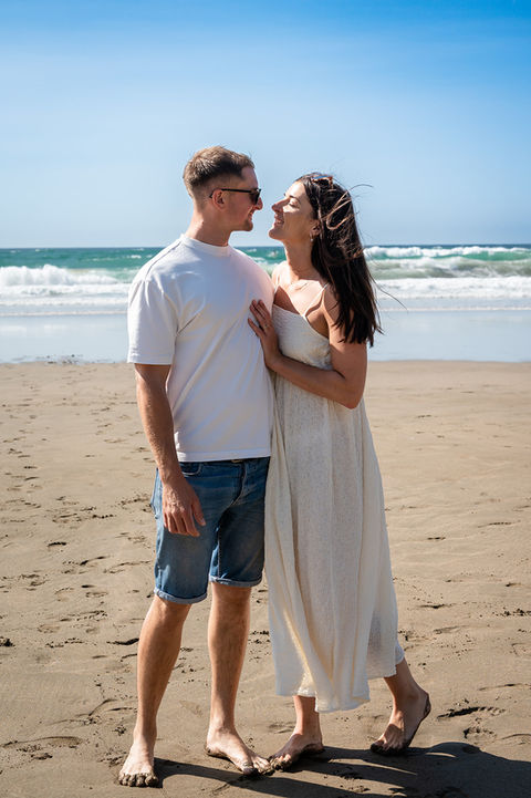 Couple standing barefoot on a sunny Cornwall beach, smiling at each other with waves in the background.